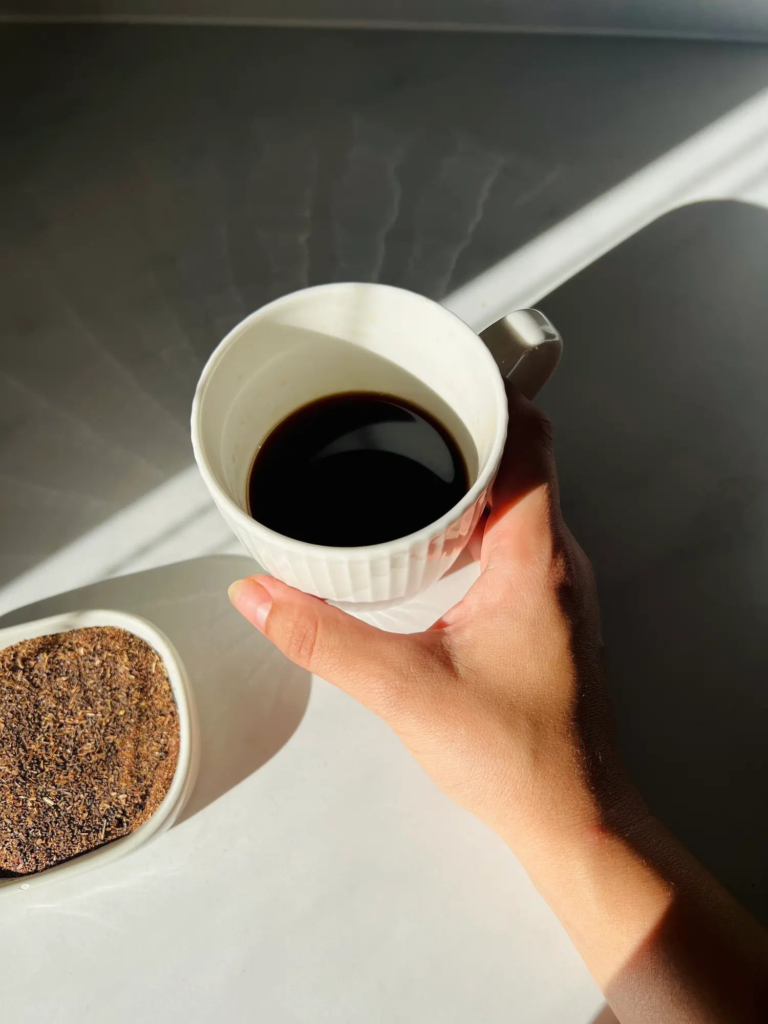 Hand holding a white cup of Karobe roasted carob drink next to a bowl of carob powder, highlighting the rich color and coffee-like appearance, surrounded by carob beans.