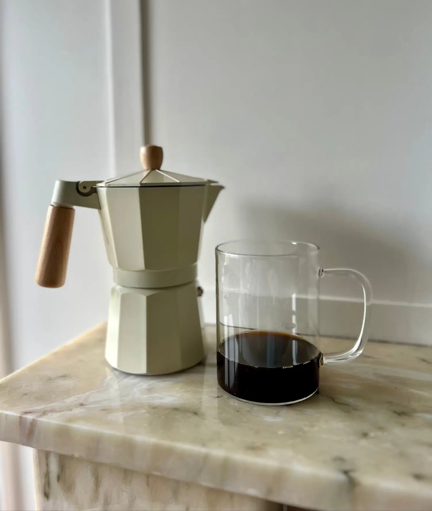 Moka pot with a glass mug filled with karobe on a marble surface.